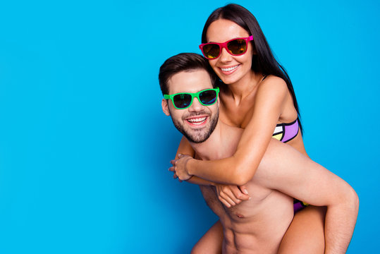 Close up of joyful couple, man in sunglasses and yellow shorts carrying on back happy woman with dark hair and looking at camera. Summer holidays concept isolated on blue background with copysapce