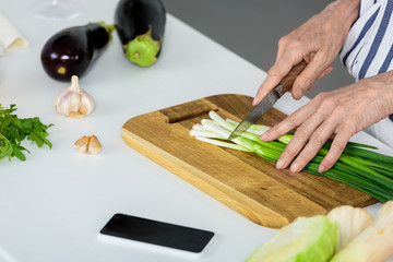 cropped image of grey hair woman cutting green onion on wooden board in kitchen