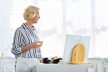 side view of attractive grey hair woman holding glass of wine in kitchen and looking away