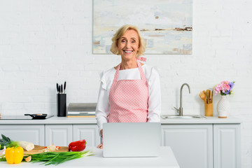 smiling attractive senior woman using laptop for cooking in kitchen and looking at camera