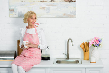 attractive senior woman sitting on kitchen counter with cup of coffee and looking away
