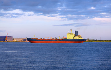 The oil products tanker is anchored in a port. The huge ice breaker is drifting near the shore in the background of a dramatic sky.