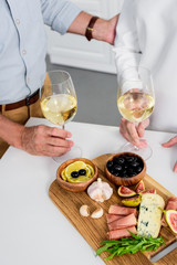 cropped shot of senior couple holding glasses of wine while enjoying delicious snacks at home