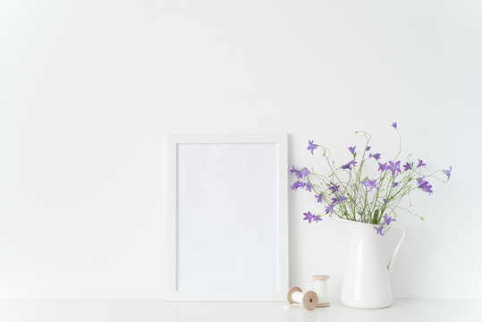 Cute white portrait frame mockup with summtr lilac wild flowers in jug, coils and tapes near wall on white background. Empty frame mock up for presentation design. Template framing for interior art.