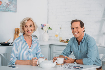 happy senior couple smiling at camera while drinking tea together at home