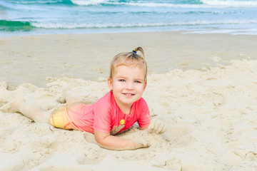 Portrait of emotional adorable toddler girl in sun protecting swimming suit lying on the sand on the beach and looking at camera. Family vacation, traveling concept. Selective focus, copy space.