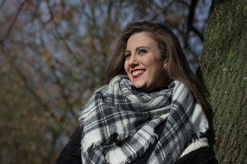 Happy young woman with squares pattern scarf leans on tree trunk on autumn season day in the park. Portrait of beautiful smiling girl on fall fashion style