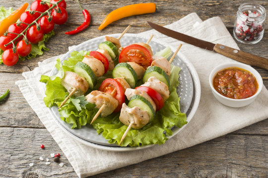 Traditional chicken kebab on wooden skewers with tomatoes, cucumbers and fresh herbs on a plate.