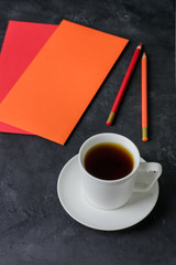 Obraz na płótnie Canvas A cup of tea, colored envelopes (orange and red) and pencils on a dark table