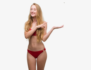 Blonde teenager woman wearing red bikini amazed and smiling to the camera while presenting with hand and pointing with finger.