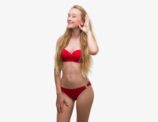 Obraz premium Blonde teenager woman wearing red bikini smiling with hand over ear listening an hearing to rumor or gossip. Deafness concept.