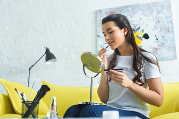 Fototapeta na wymiar young woman applying lipstick while doing makeup on sofa at home