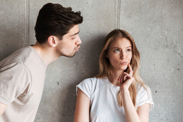 Photo of outraged doubtful woman hesitating while handsome man kissing her cheek, isolated over gray background