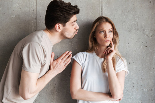 Image of outraged sassy woman expressing unwillingness while brunette man kissing her cheek, isolated over concrete wall