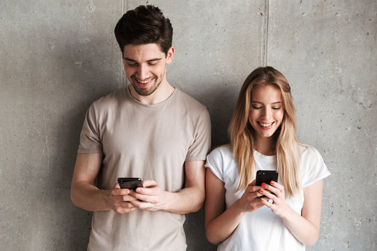 Photo of positive people man and woman smiling while both using mobile phones, isolated over concrete gray wall indoor