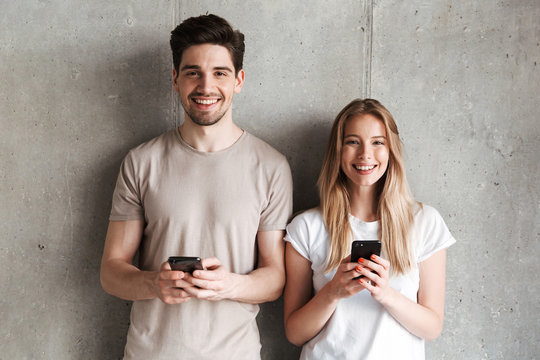 Modern sociable people man and woman smiling while both holding smartphones, isolated over concrete gray wall indoor