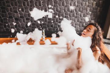 Happy woman playing with foam in big bath with wooden edge. Relaxation in spa