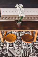 Modern wooden furniture in cafe on floor with black and white ornament tile. Two chairs, table, sofa, orchid flower
