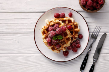 freshly baked belgian waffles with raspberries on plate with cutlery on white wooden table