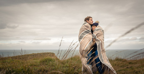 Portrait of young beautiful couple embracing under blanket in a cold day with sea and dark cloudy...