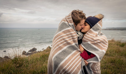 Fototapeta premium Portrait of young beautiful couple kissing under blanket in a cold day with sea and dark cloudy sky on the background
