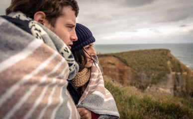 Closeup of young beautiful couple under blanket in a cold day looking the sea with the dark cloudy sky on the background