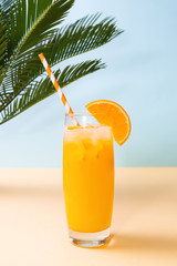 Glass of fresh orange juice and green palm leaves on pastel background. Copy space. Pop art design, minimal summer concept.