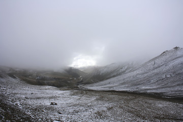 a foggy view of a mountain pass; sunlight makes its way through a misty veil; break in the clouds; mountain serpentine highway and grazing sheep in the distance; the first snow in the highlands, Tibet