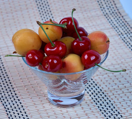 Lots of ripe fresh apricots and cherries in a glass vase on a bamboo napkin.
