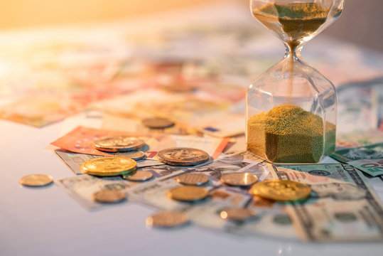 Sand running through the shape of hourglass on table with banknotes and coins of international currency. Time investment and retirement saving. Urgency countdown timer for business deadline concept