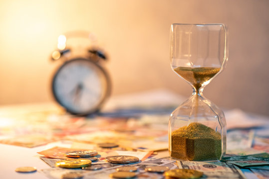 Sand running through the shape of hourglass on table with banknotes and coins of international currency. Time investment and retirement saving. Urgency countdown timer for business deadline concept