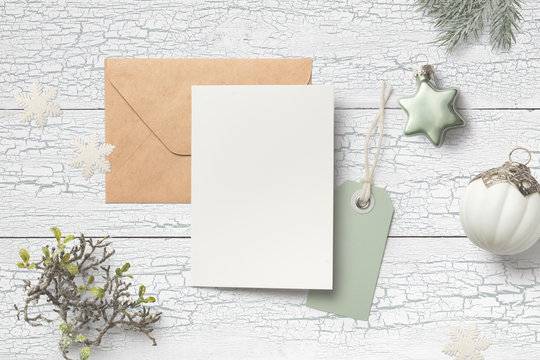 minimalist christmas card mock up (portrait format) with green fir twigs, baubles, card, envelope, hangtag and glitter snowflakes on a white wooden background