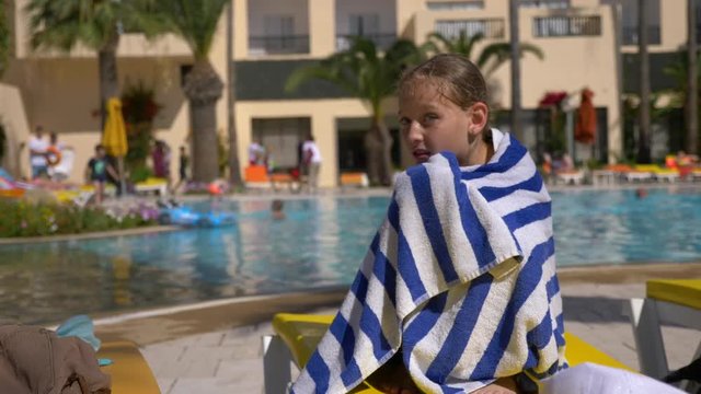 Young wet girl wrapped in towel sits on sunbed near swimming pool