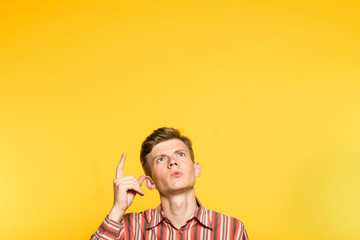 amazed impressed astonished man looking and pointing up with a finger. portrait of a young guy on yellow background popping up or peeking out from the bottom. free space for advertisement. - obrazy, fototapety, plakaty