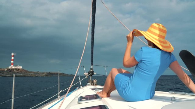 Woman in a yellow hat and blue dress girl rests aboard a yacht near the lighthouse on summer season at ocean