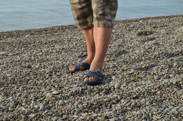 men's legs in flip-flops and shorts, a man standing on the beach on a pebble beach