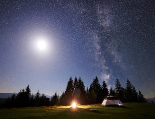 Camping site in mountain valley at night. Male hiker standing near tourist tent at campfire under night blue starry sky with Milky way and bright full moon. Outdoor activity and traveling concept