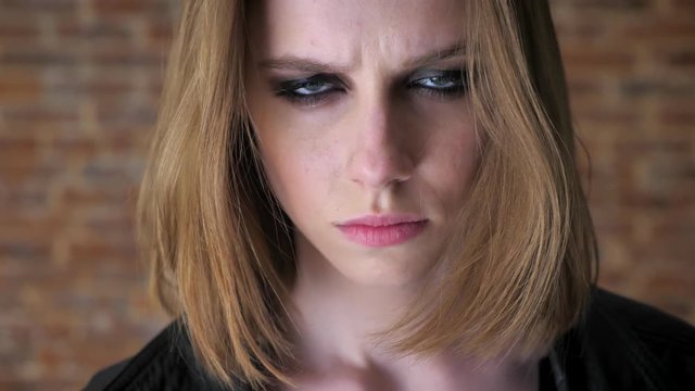 Young sexy disgusted girl with smoky eyes is watching at camera, calming down, brick background, blurred background