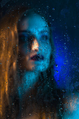 Art style portrait in blue tones of a lonely beautiful sad model girl behind a window glass, over which rain drops down. Artistic blur on model face.