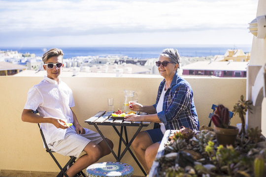 happy leisure activity on the terrace rooftop having breakfast with smiles and happiness for grandmother and teenager family caucasian people. ocean and buildings view, outdoor coule together