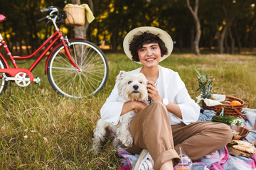Smiling girl in hat hugging little dog happily looking in camera spending time on picnic in park with bicycle on background