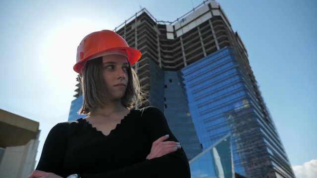 Young strong girl builder with hemlet is standing with arms crossed in daytime in summer, building concept, urban concept, building on background, bottom view