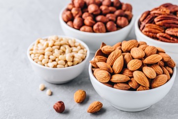 Almonds, pecans, pine nuts and hazelnuts in white bowls on grey background. Nuts mix.