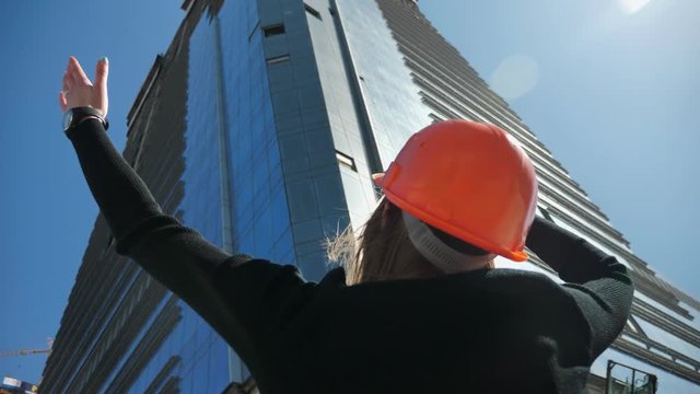 Young girl builder with hemlet is watching at building, showing greatness of it in daytime in summer, building concept, urban concept, bottom view