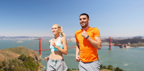 fitness, sport and healthy lifestyle concept - smiling couple with heart-rate watch running over golden gate bridge in san francisco bay background