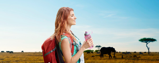 adventure, travel, tourism, hike and people concept - smiling young woman with backpack and bottle of water over animals in african savannah background