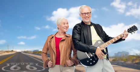 rock music, old age and travel concept - happy senior couple with electric guitar over us route 66 background