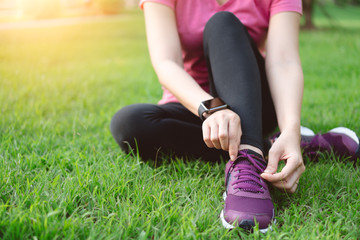 Young woman runner tying shoelaces in lawn outdoors. Fitness female runner wear running shoes ready for workout and jogging in park at summer with smart watch heart rate monitor. Healthy lifestyle.