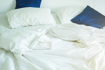 white pillow and dark blue on bed and with wrinkle messy blanket in bedroom, from sleeping in a long night, an unmade bed in hotel bedroom with white blanket.