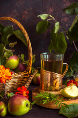 Apple tea in a glass goblet and a decanter and a basket with ripe apples on a wooden table in...